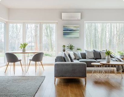 White room with large windows and a heat pump mounted on the wall. The room has grey couches and mat