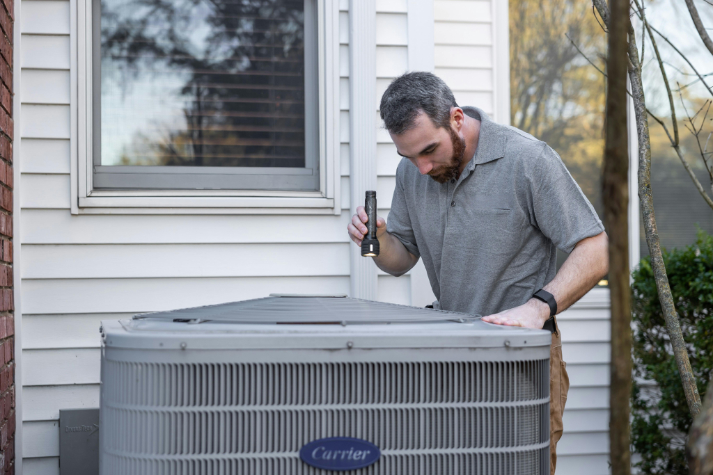 HVAC technician checking on a condenser unit with a flashlight.
